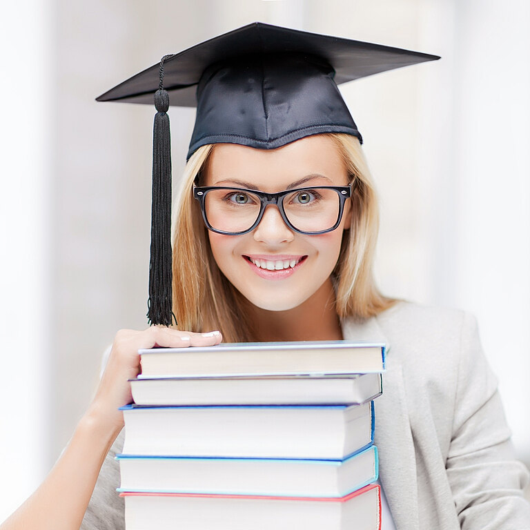 Mädchen hat einen Doktorhut auf, lacht und hat vor sich einen Stapel Bücher