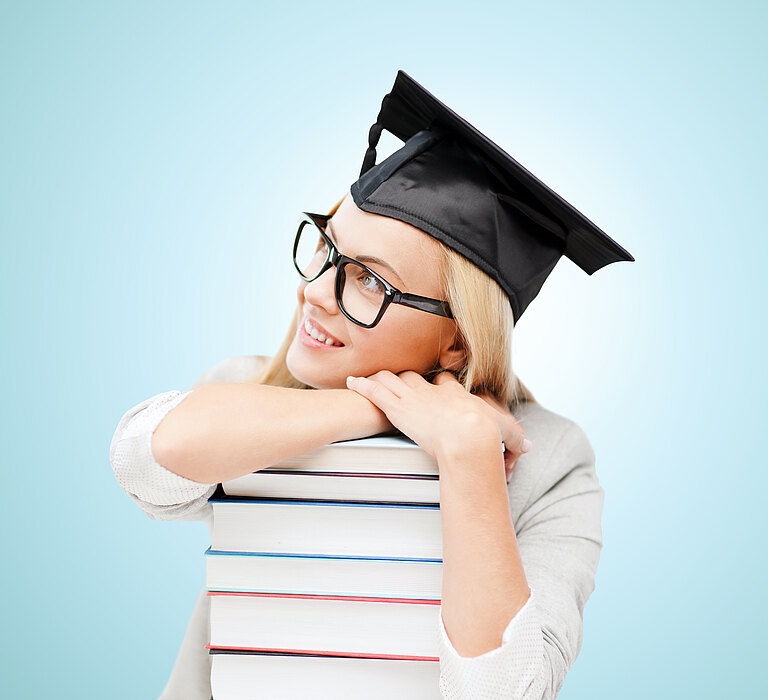 Mädchen hat vor sich einen Stapel Bücher und trägt einen Doktorhut. Sie lacht zufrieden.